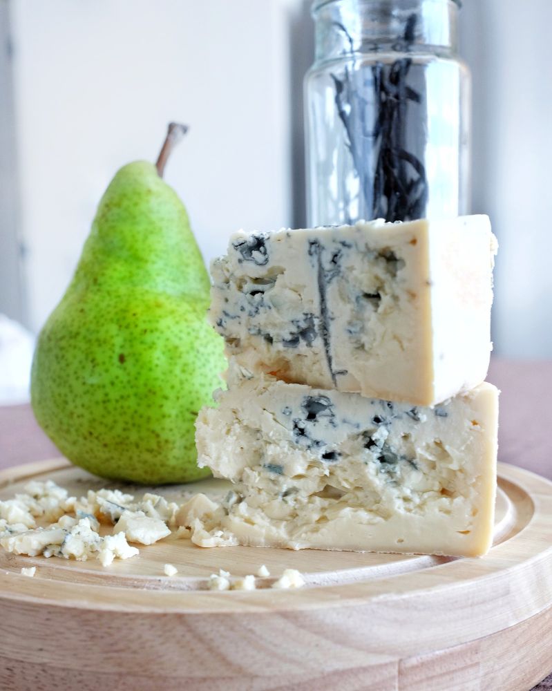 2 wedges of a marble blue cheese stacked on top of each other on a round wooden cheese board. There's a juicy looking green pear standing upright behind the cheese