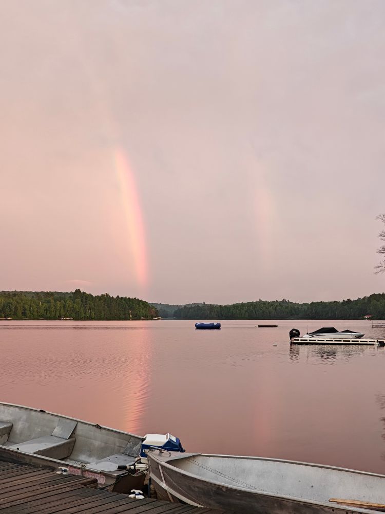 Double rainbow in a grey sky after a rain storm, reflected in the glassy still water of Oxtongue Lake, Muskoka. In the foreground are two aluminum boats tied to a dock.