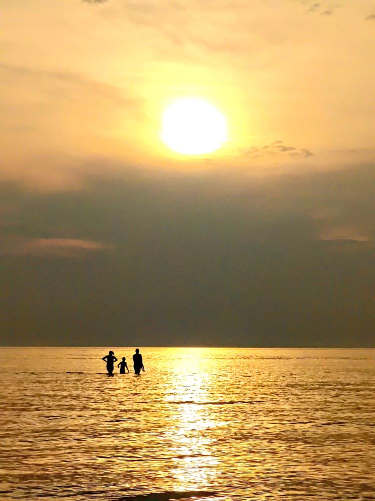 The silhouettes of two adults and a child are standing in a lake of golden water under a setting sun that is just above a bank of dark clouds
