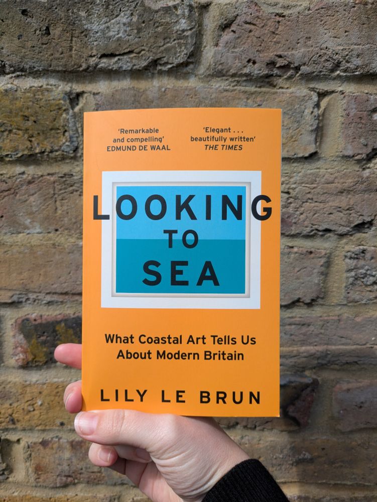 A paperback book held up against a brick wall. LOOKING TO SEA: WHAT COASTAL ART TELLS US ABOUT MODERN BRITAIN, by Lily Le Brun. "Remarkable and compelling" Edmund de Waal, "Elegant... Beautifully written" The Times 