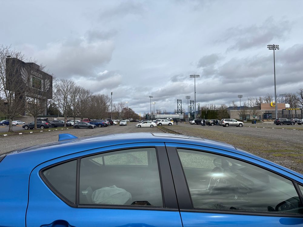 A blue car parked at an extreme end of a parking lot, with a baseball field barely visible in the distance 