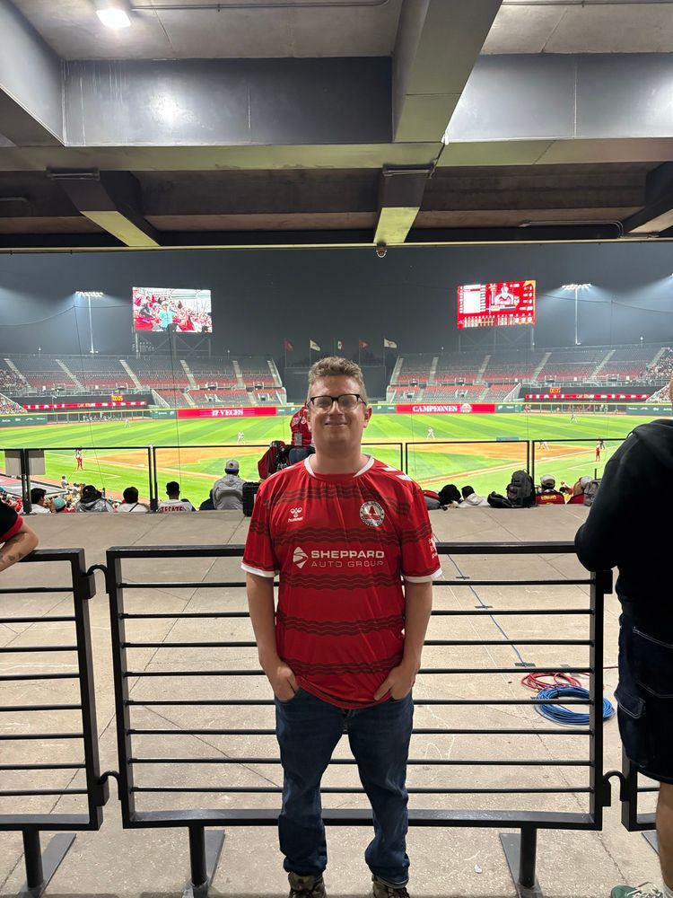 A man in a red soccer jersey in a baseball stadium 