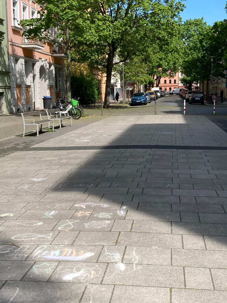 Verkehrsberuhigter Straßenabschnitt vor einer Autos die Durchfahrt verhindernden Poller-Reihe. Auf dem Boden mitten auf der Straße Kreide-Malereien von Kindern