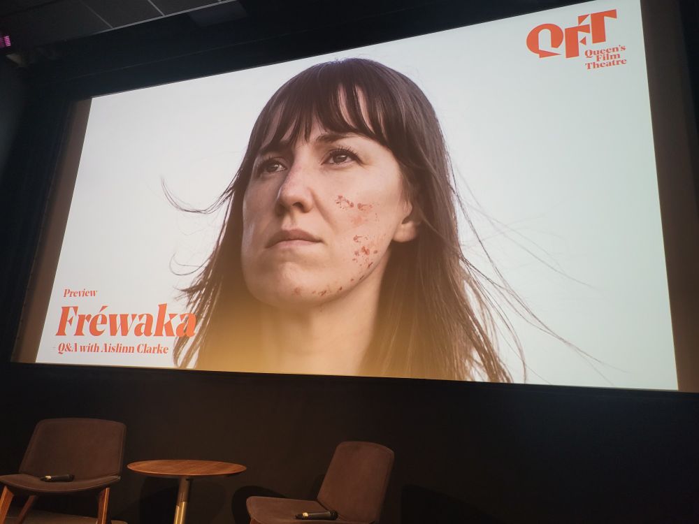 A photo taken from the film Fréwaka, projected on a cinema screen at Queens Film Theatre, with a table and chairs in the foreground. 