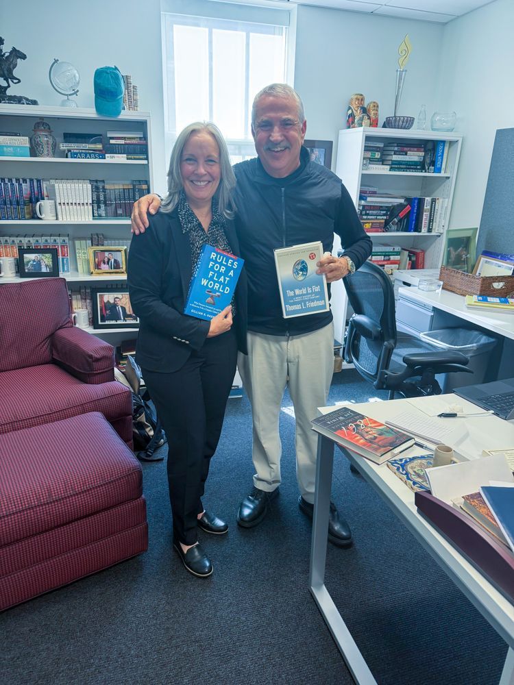 Gillian Hadfield and Thomas Friedman stand together smiling in an office, each holding their respective books - Hadfield holds 'Rules for a Flat World' and Friedman holds 'The World is Flat.' 
