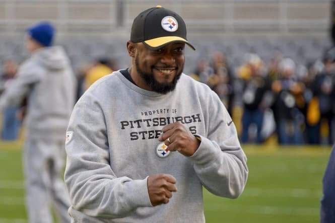 Steelers Head Coach Mike Tomlin wearing a gray Pittsburgh Steelers sweatshirt and black Steelers baseball hat, standing and smiling while making two fists  
