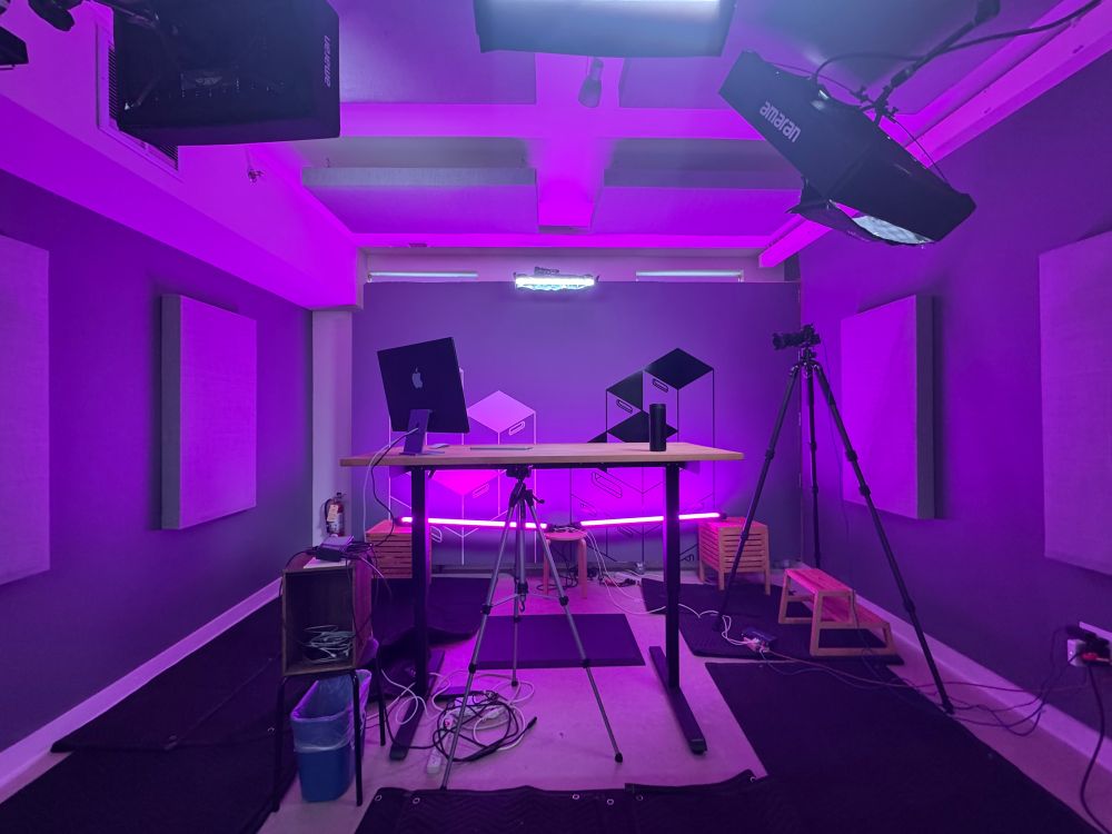 the interior room of a Baltimore row house which has been converted to a small recording studio for creating video content. the room has a standing desk in the center and is lit in a rich purple hue. an iMac sits on the standing desk, while various cameras and pieces of camera equipment surround the desk. sound baffling materials are visible on the ceiling, walls, and floor