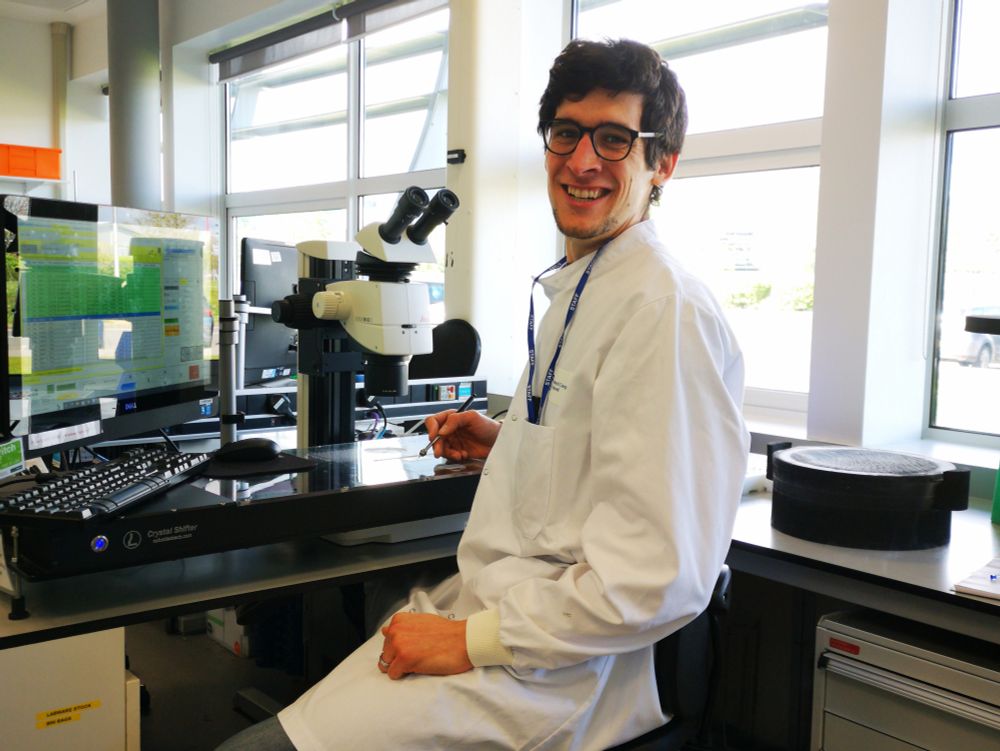 Smiling Hans in a lab coat sitting with a crystal-harvesting wand in front of a Crystal Shifter robot with a microscope and a foam bucket of liquid nitrogen.