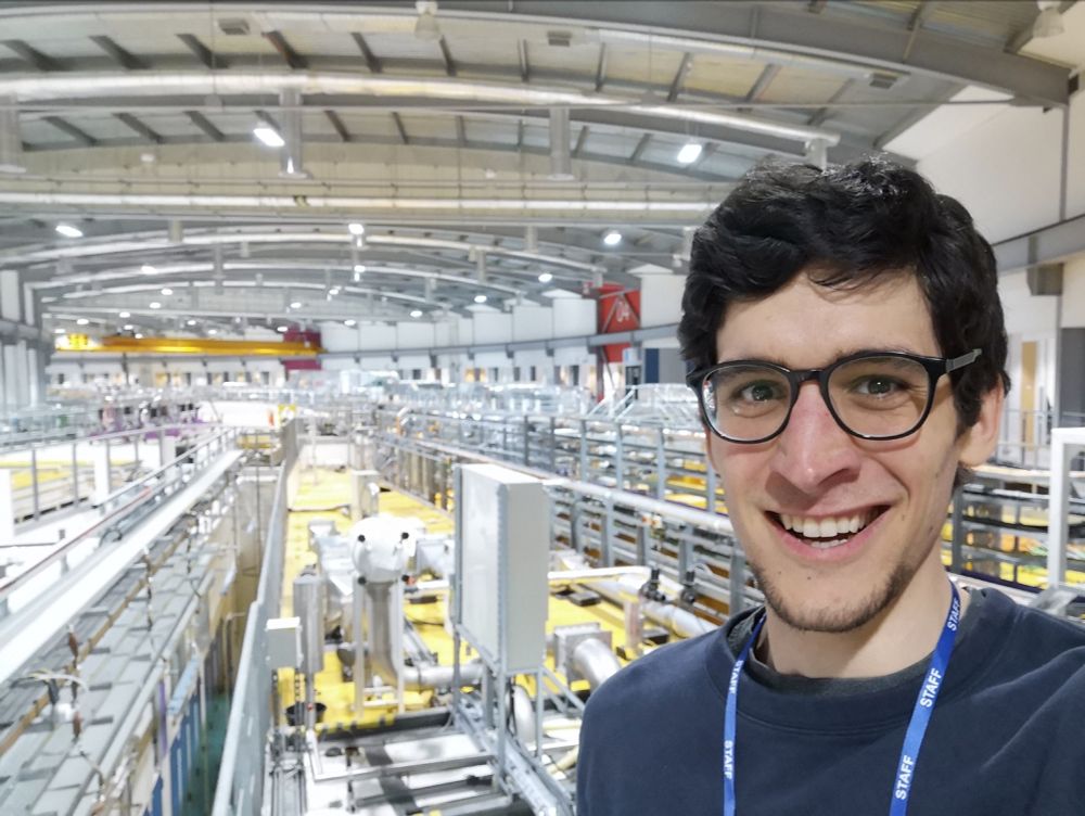 A smiling Hans with a staff lanyard, standing inside the experimental hall of Diamond Light Source, the UK national synchrotron. There are the roofs of a couple beamlines and a yellow crane at the back. The curved wall has zone 4 indicated in red.