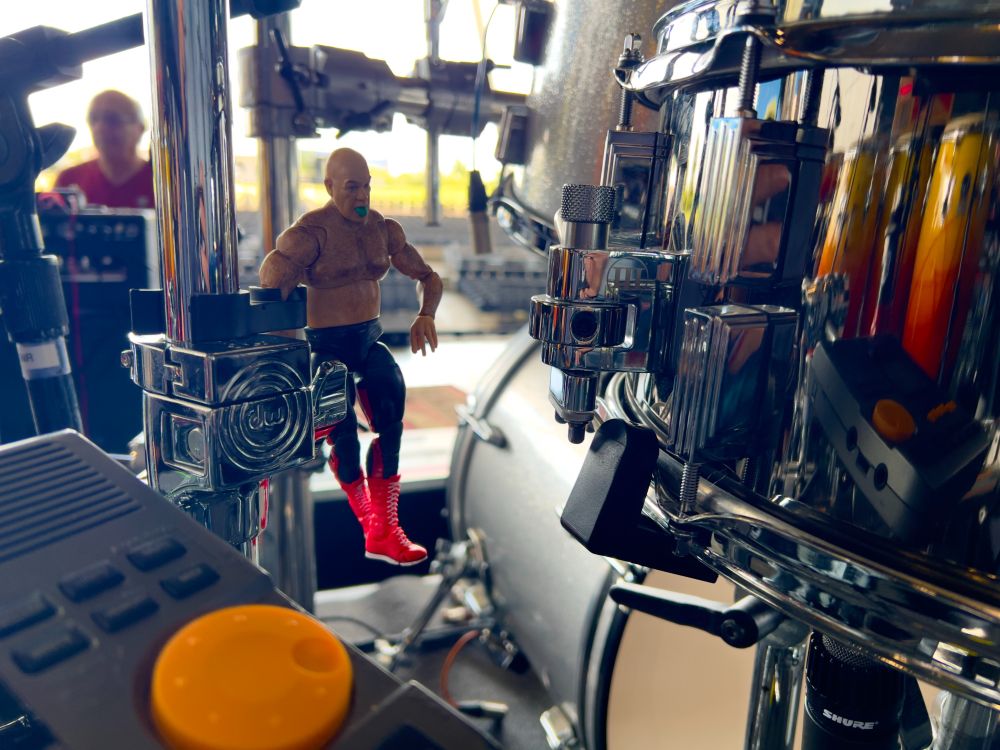 An wrestling action figure attached to a drum stand with amphitheater seating in the background.