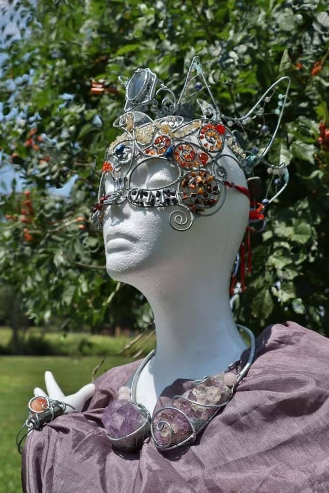 A set of handmade wire jewelry displayed on a mannequin with one hand raised and wrapped in shiny mauve fabric. The set consists of a flame-themed masquerade mask with a variety of gemstone beads, a wave-themed banana clip crown fronted by a large labradorite with a variety of other stones, a collar with two large chunks of amethyst geode and a few round rose quartz beads, and a cuff bracelet with carnelian, pyrite, and labradorite. 