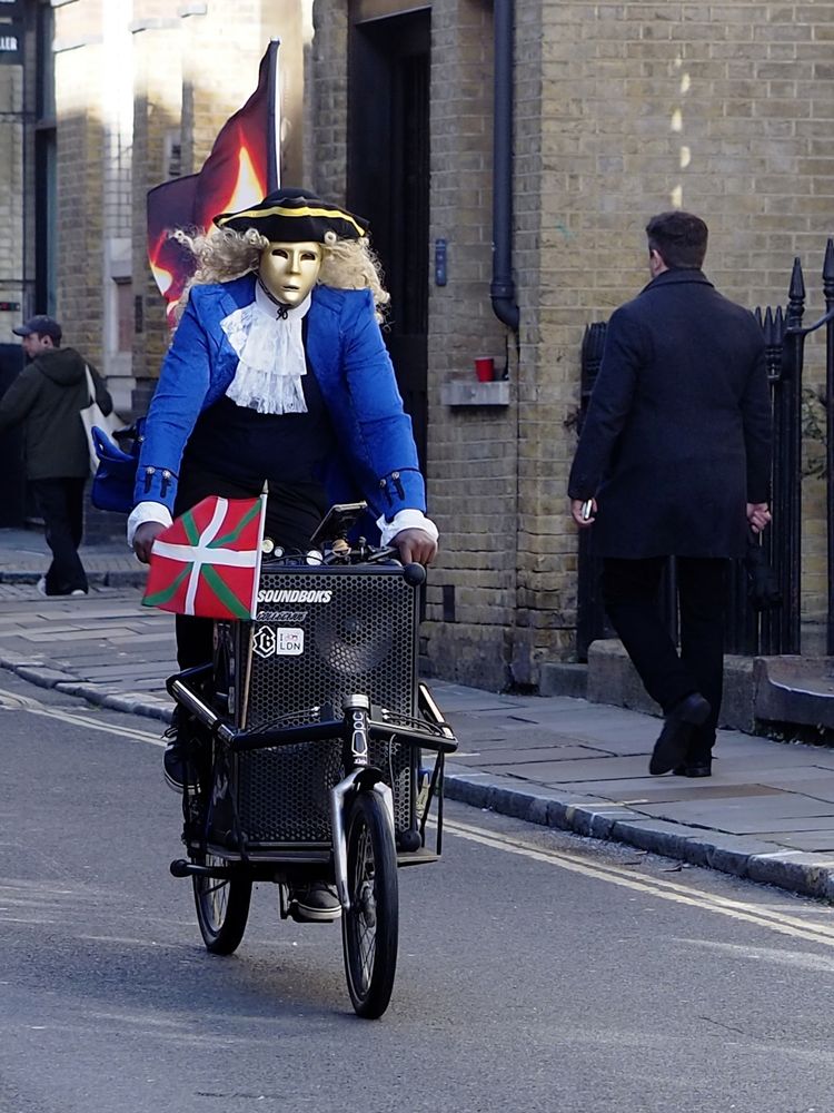 Man dressed like an escapee from Amadeus in a blue coat, white jabot, black tricornered hat with gold trim. He's riding a black electric cargo bike with a sound system which is blasting a disco version of Mozart. His face is concealed by a gold mask. A Basque flag flies from the front of the bike 