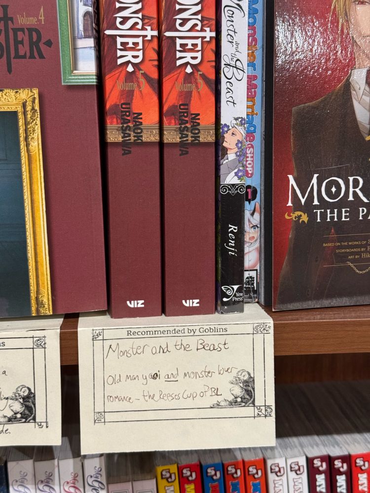 A wooden bookshelf with several manga books, and asmall handwritten paper sign. The sign reads: monster and the beast, old man yaoi and monster lover romance hyphen the Reese's cup of BL