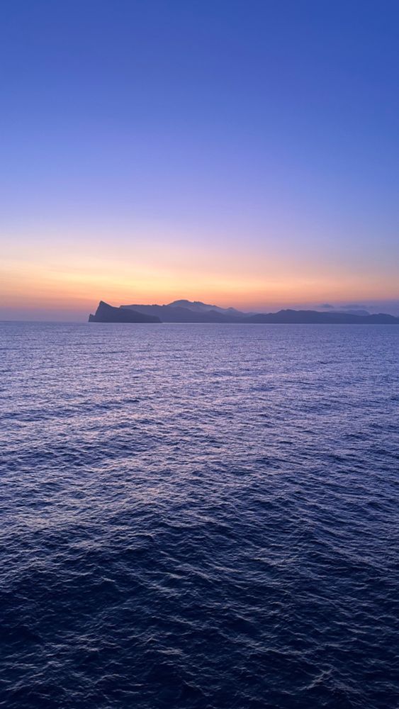 a band of gold and orange dawn light bisects the top third of a purple-toned photograph of sea and sky with the hazy outline of Mallorca’s coast in the distance