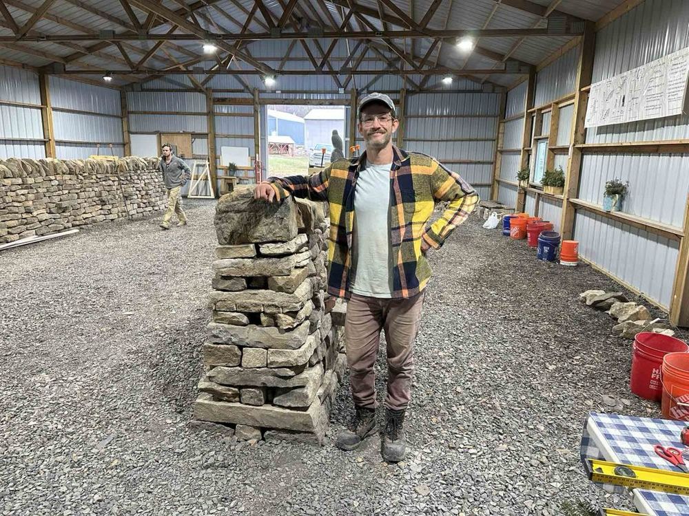 Ian leaning against the cheek end of a dry stone wall he built in 7.5 hours as part of a practice test for a masonry certification 