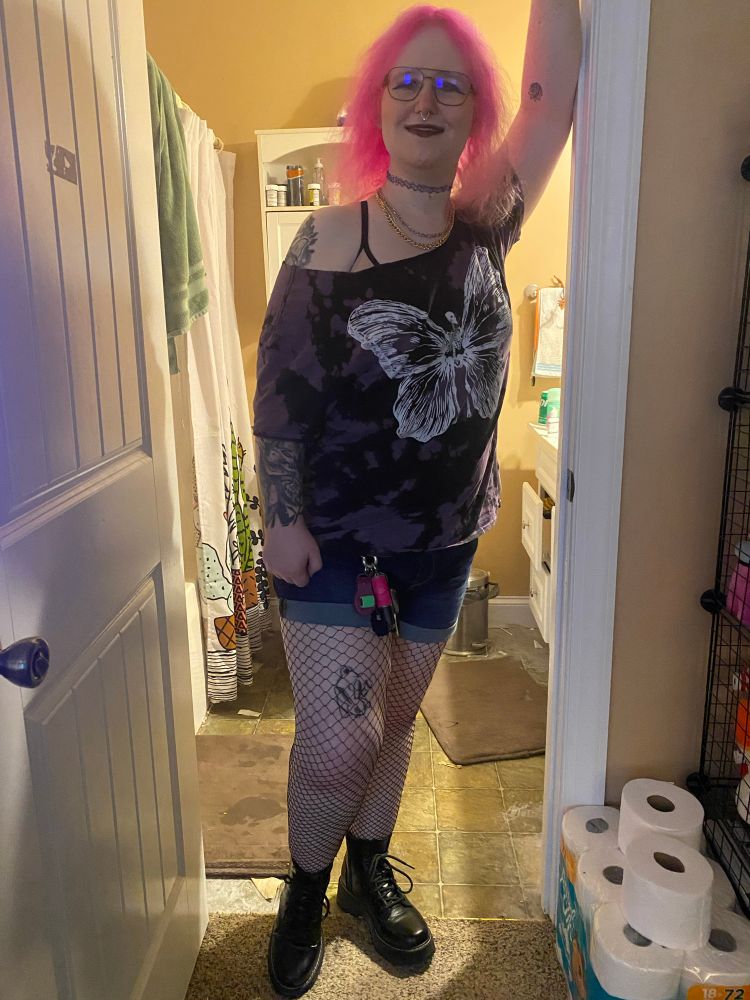 Picture of myself with my pink hair down while I wear blush and makeup along with purple lipstick and eyeshadow. Wearing a purple tie dye shirt with a shoulder cut. The graphic on it deprives a skeleton with butterfly wings. Full body photo reveals bottom with fishnets and boots along with jean shorts. A carabiner holds several keys, key chains and a clicker. A rabbit tattoo is visible on my left thigh right above my knee.