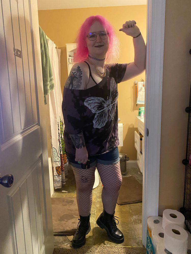 Picture of myself with my pink hair down while I wear blush and makeup along with purple lipstick and eyeshadow. Wearing a purple tie dye shirt with a shoulder cut. The graphic on it deprives a skeleton with butterfly wings. Full body photo reveals bottom with fishnets and boots along with jean shorts. A carabiner holds several keys, key chains and a clicker. A rabbit tattoo is visible on my left thigh right above my knee. Same framing just showing off more of my Mothman tattoo 
