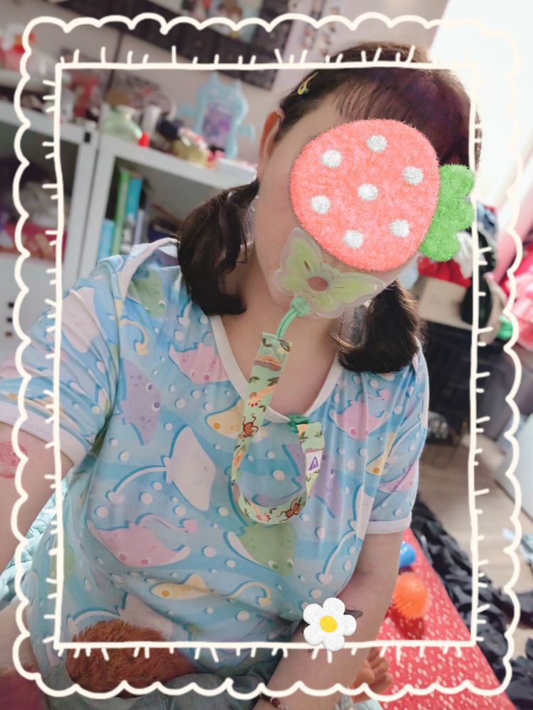 A picture of a white person with brown hair in low pigtails. They are wearing an adult size baby onesie in light blue with colorful stingrays on it and are using a green butterfly shaped pacifier. Their face is censored with a strawberry and their tattoo is censored with a flower