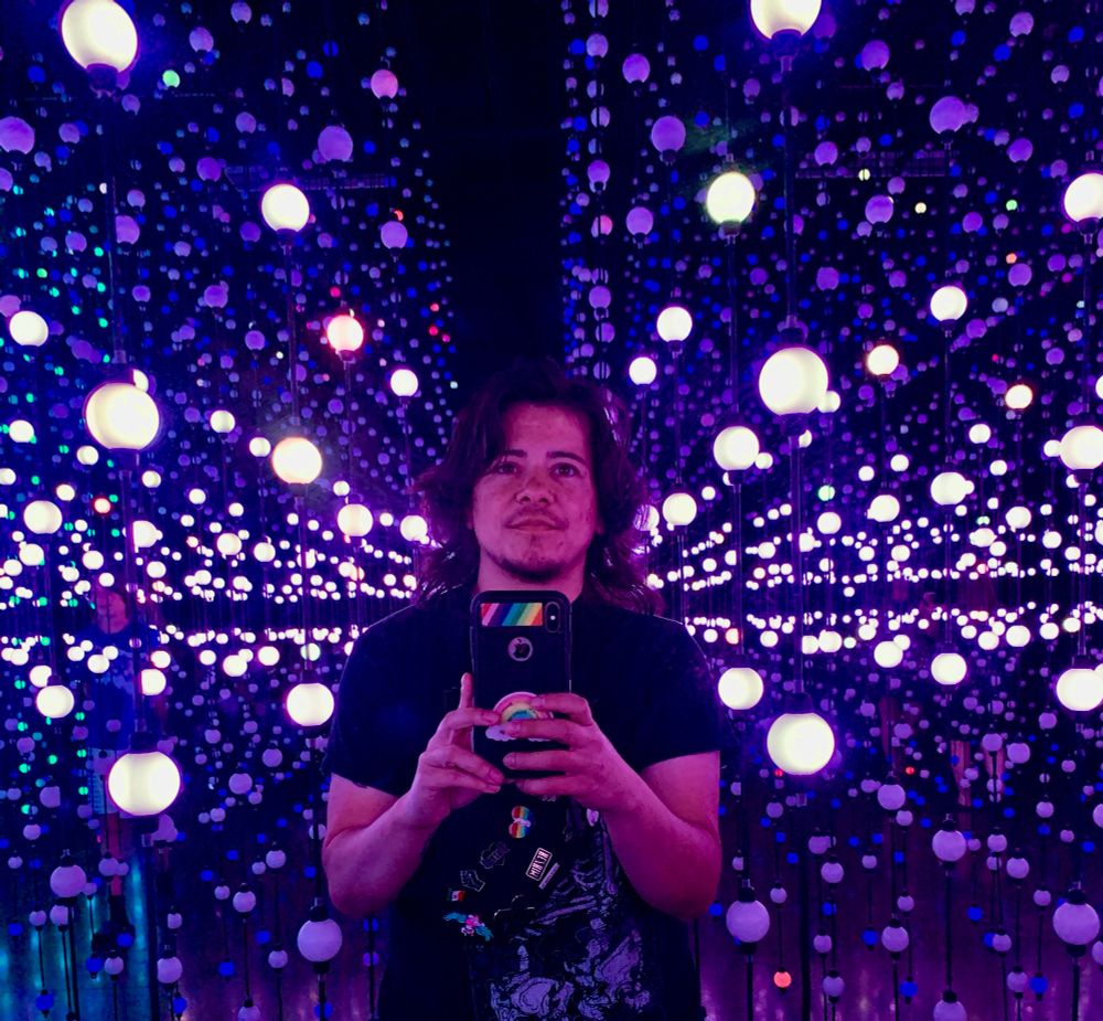 Leon taking a selfie in the exhibit, Matrix, an imermersive piece that is hundreds of strings of orbs that light up orchestrating with music in a dark room. The orbs are lit a shade of blues and purples.