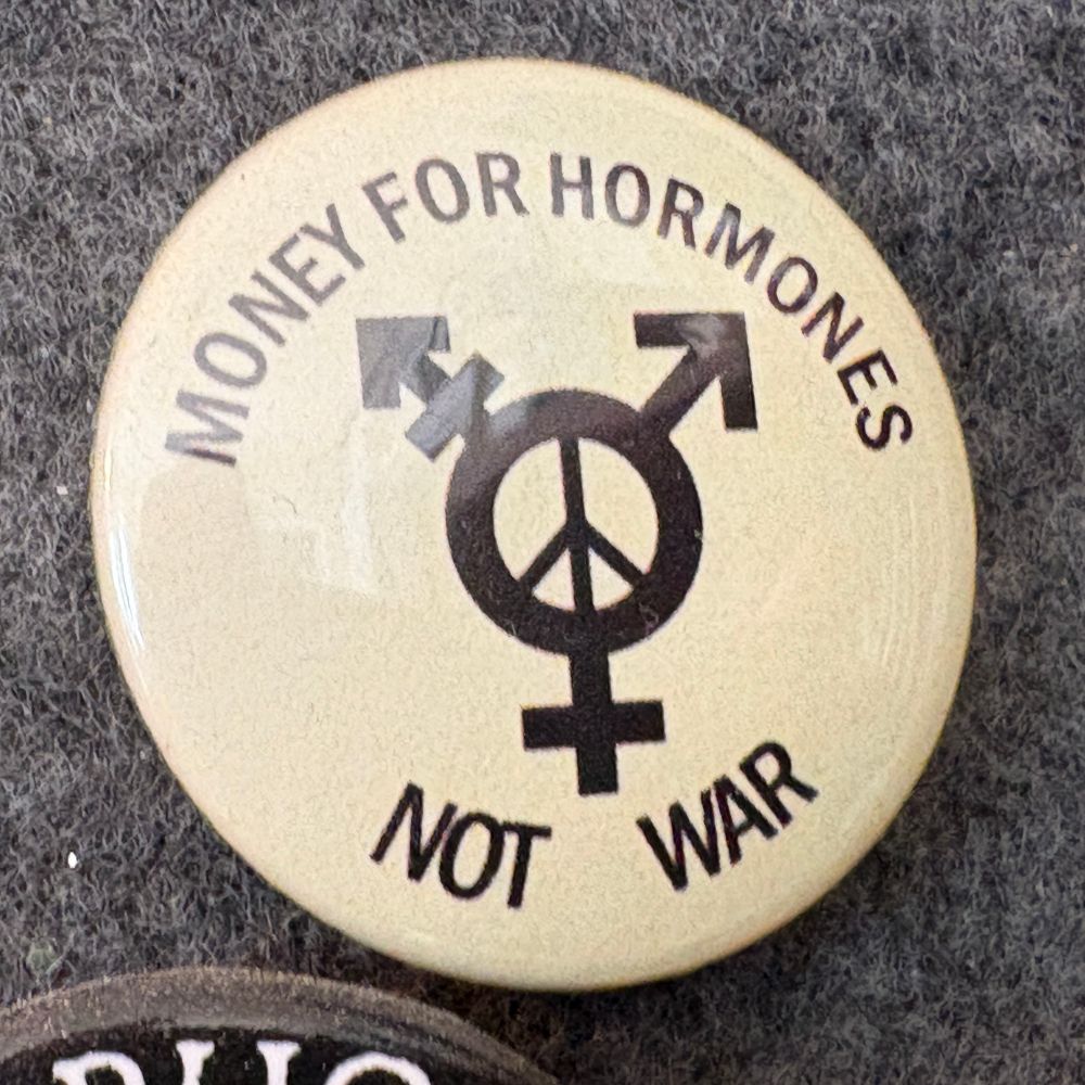 A pinback button. It shows the transgender “trine” combining three sex symbols, with a “peace sign” in the middle. Around the edge it reads, “Money for hormones, not war.”