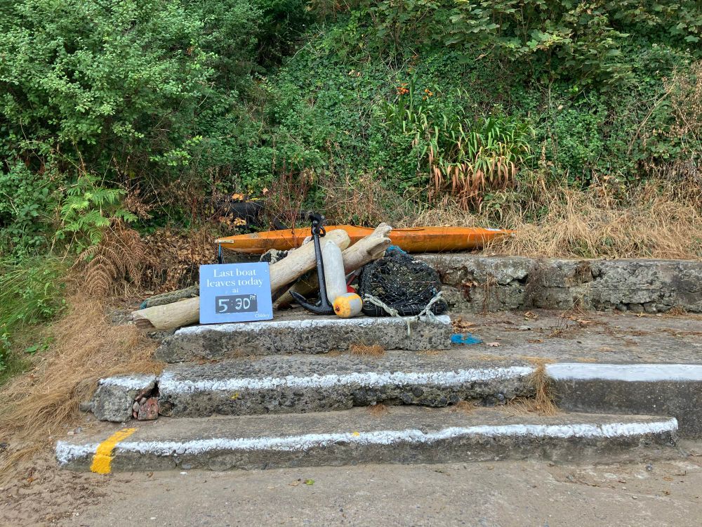 Auf Steinstufen steht ein Schild mit der Aufschrift „Last boat leaves today at 5:30“. Dahinter liegt eine Ansammlung von maritimen Gegenständen, unter anderem Strandholz und ein orangenes Kajak. 