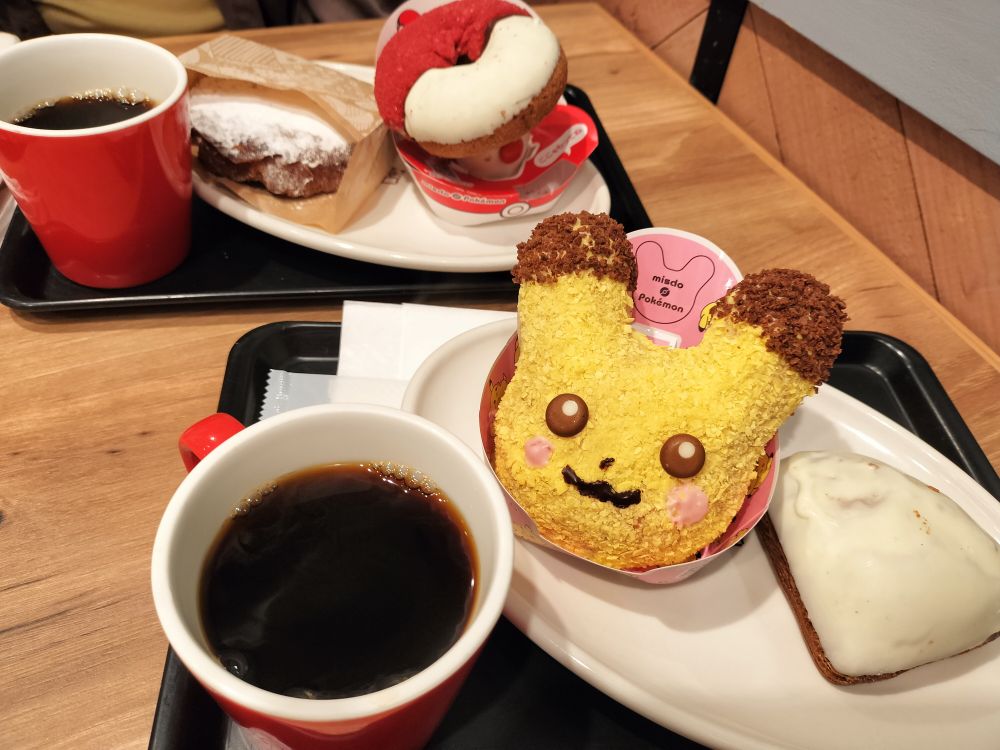 ミスタードーナツの店内。木目のテーブルに2人分のトレーがあり、それぞれにホットコーヒーとドーナツがのっている。手前のトレーにはピカチュウの顔のドーナツがあり、おいしそう。
Inside a Mister Donut. Two trays are placed on a wooden table, each holding a hot coffee and donuts. The tray in front holds a delicious-looking Pikachu-shaped donut
