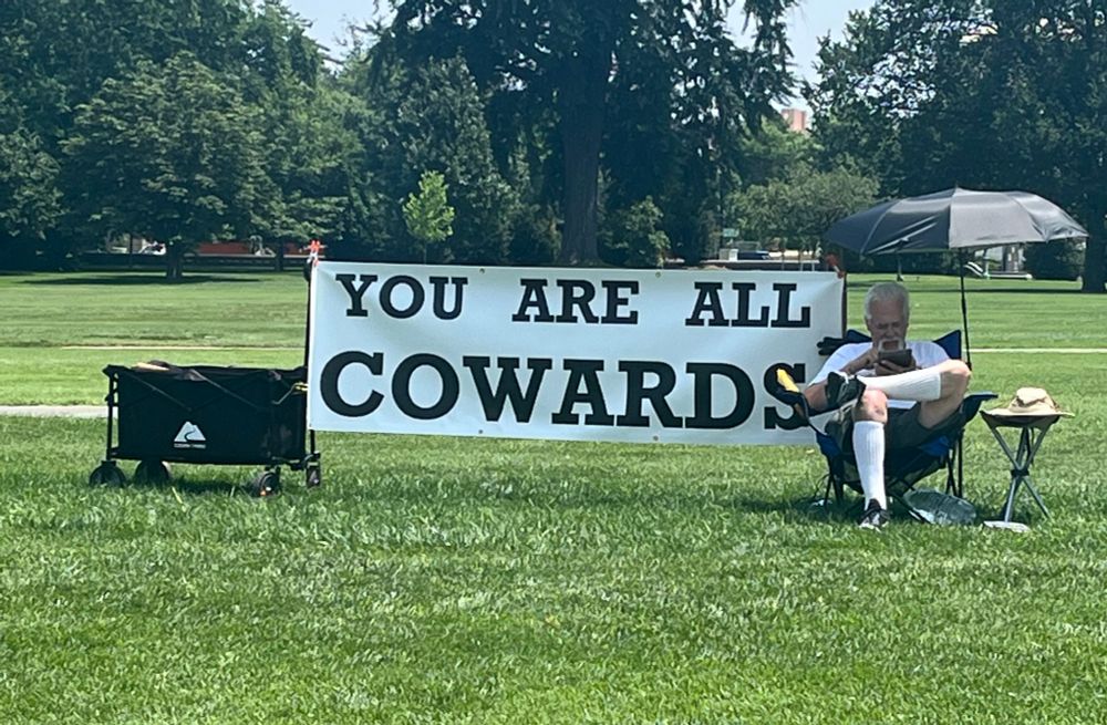 Person sitting with a banner that reads YOU ARE ALL COWARDS