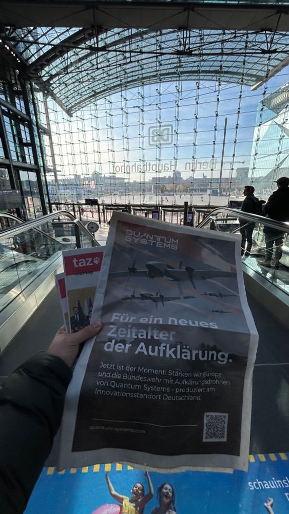Eine Person hält eine Zeitung mit einer Werbeanzeige von Quantum Systems in der Hand. Die Anzeige zeigt mehrere Drohnen im Flug und trägt die Überschrift: ‘Für ein neues Zeitalter der Aufklärung.’ Darunter steht ein Text, der die Bundeswehr und Europa zur Nutzung von Aufklärungsdrohnen aus deutscher Produktion aufruft. Im Hintergrund ist der Berliner Hauptbahnhof mit seiner großen Glasfassade zu sehen. Die Stadtlandschaft und einige Passanten sind ebenfalls im Bild.