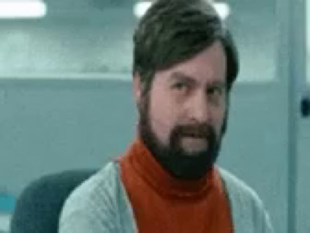 a man with a beard and a turtleneck is sitting in a chair and making a funny face .