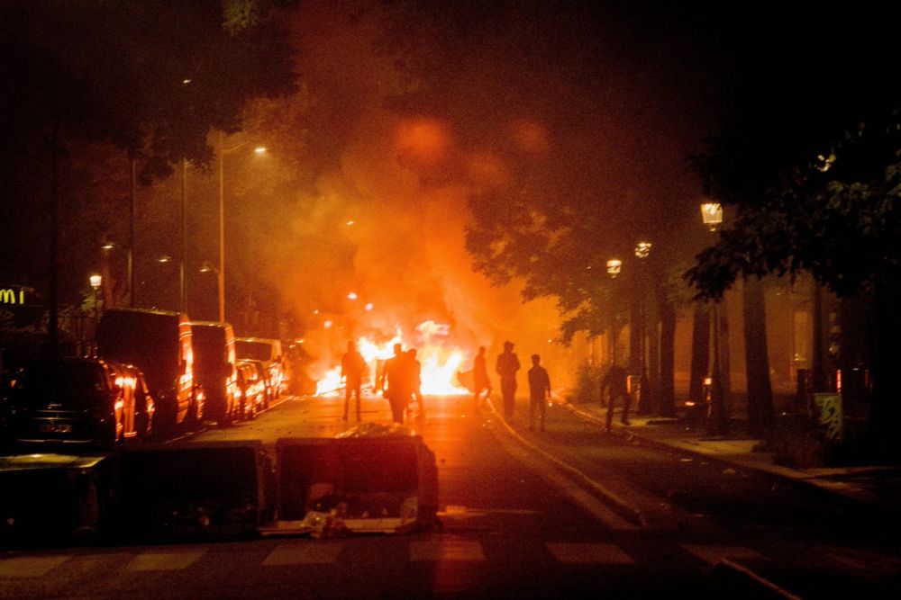 Auf dem Bild sind einige Silhouetten vor einer brennende Barrikade zu sehen. Davor legen einige umgekippte Müllcontainer zu sehen.