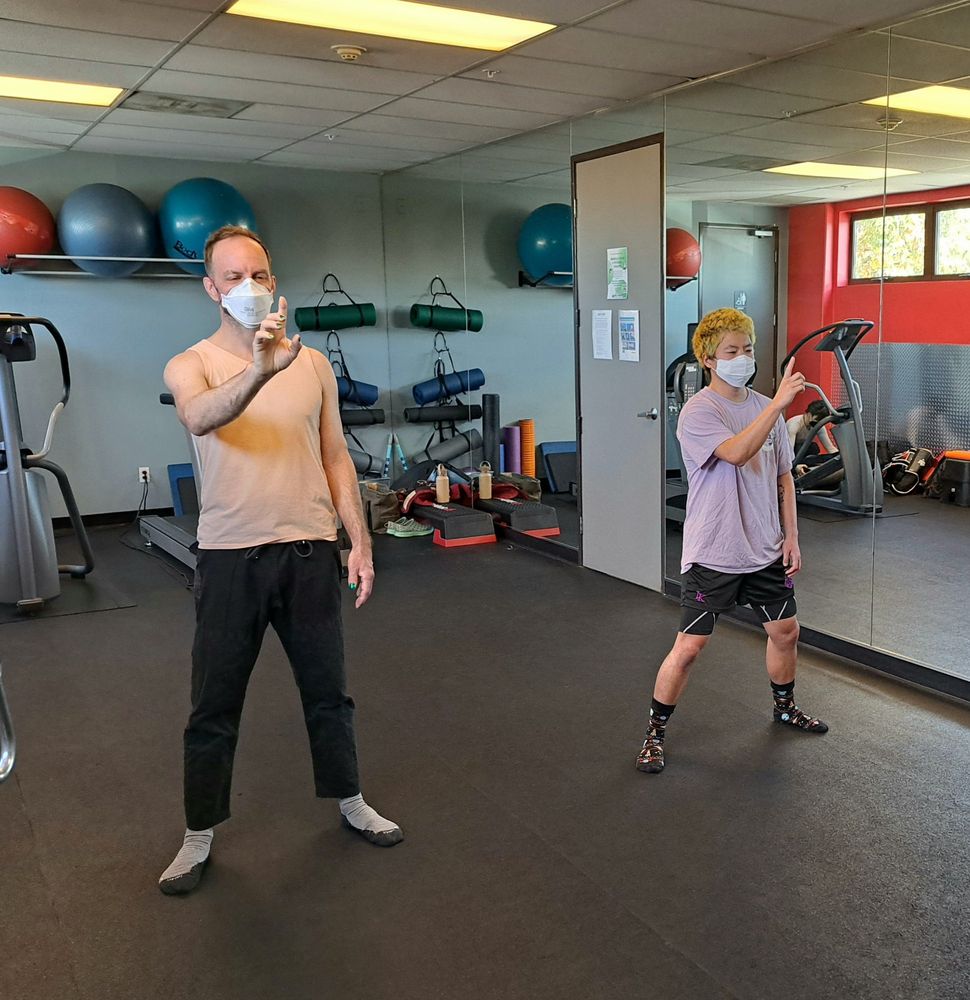 Two people wearing masks are in a gym setting with mirrored walls and exercise equipment in the background. Both are standing in slightly bent stances, staring at their index fingers.