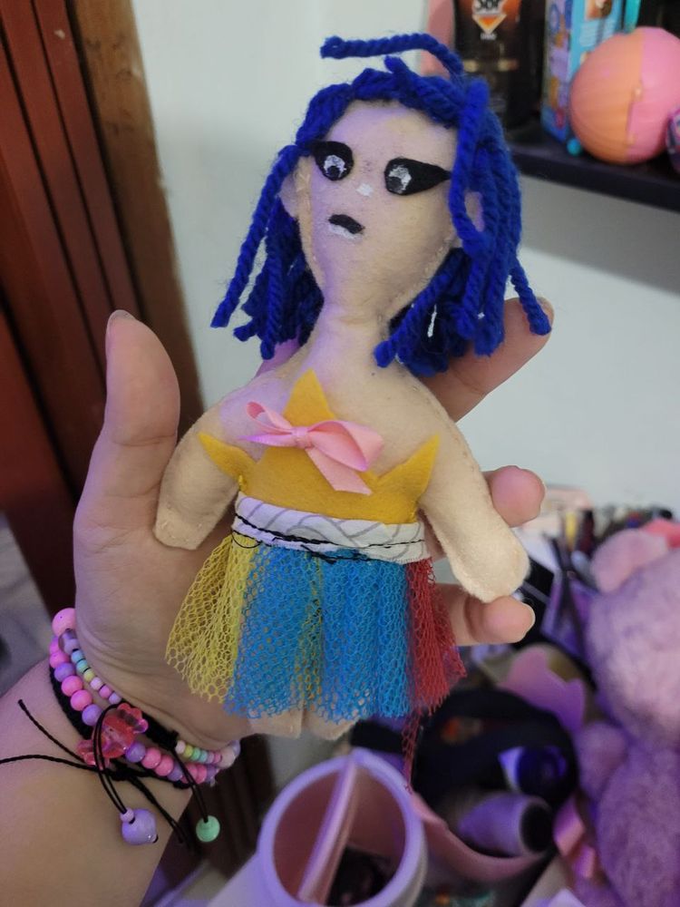Bonequinha de uma pessoa branca (cor rosa clarinho) de cabelo azul ate os ombros, olhos com delineado enorme preto, blusa em formato de coroa com lacinho rosa, a saia de filo colorido