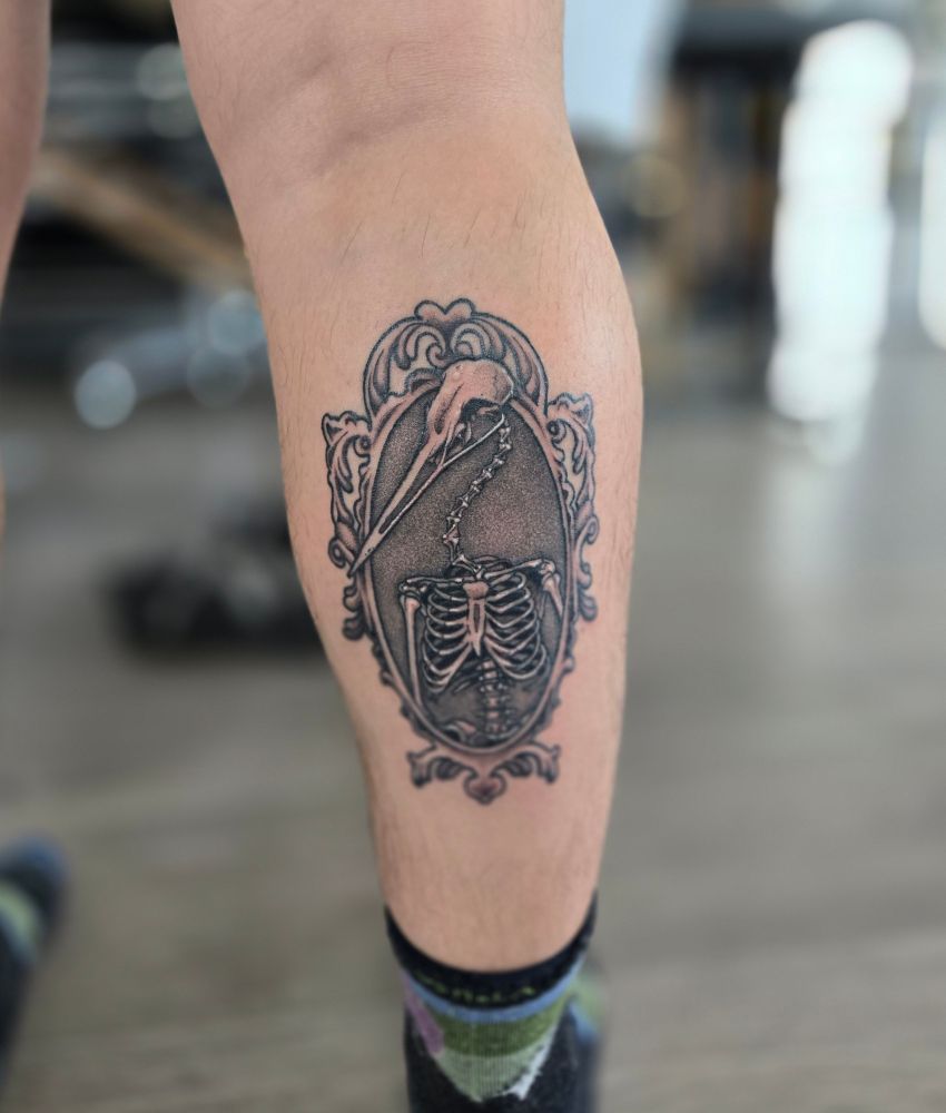 a dnd-inspired cryptid tattoo of a human skeleton with the skull of a sandhill crane. the tattoo has a soft grey background and is accented by an ornate vintage frame. 
