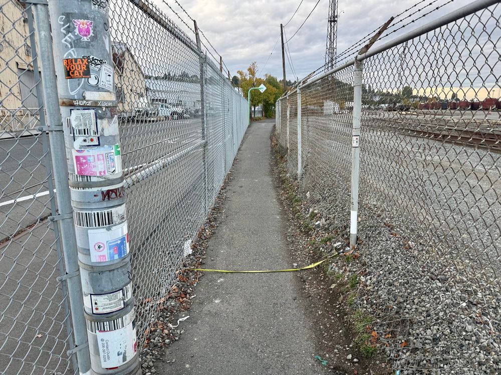 A tape measure is laid across a pinch point on a trail made narrow by chain link fencing on each side 