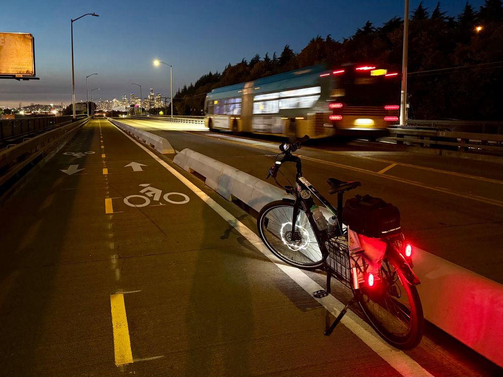 My e-bike sits in a 2-way bike lane protected by concrete mini jersey barriers as a King County Metro bus passes by with a view of the Seattle skyline at night 