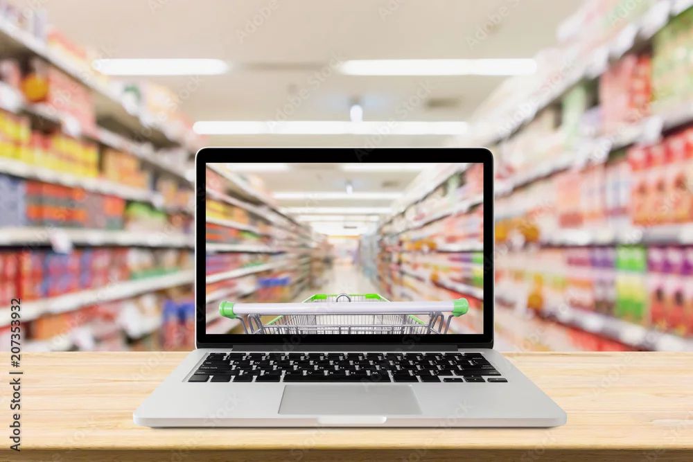 a composited stock image from adobe stock (file #207353932) depicting an open laptop on a table in the middle of a heavily blurred grocery aisle. on the laptop's screen is that same blurry grocery aisle, but with a shopping cart in the foreground creating a sort of POV image of pushing a cart down the aisle.