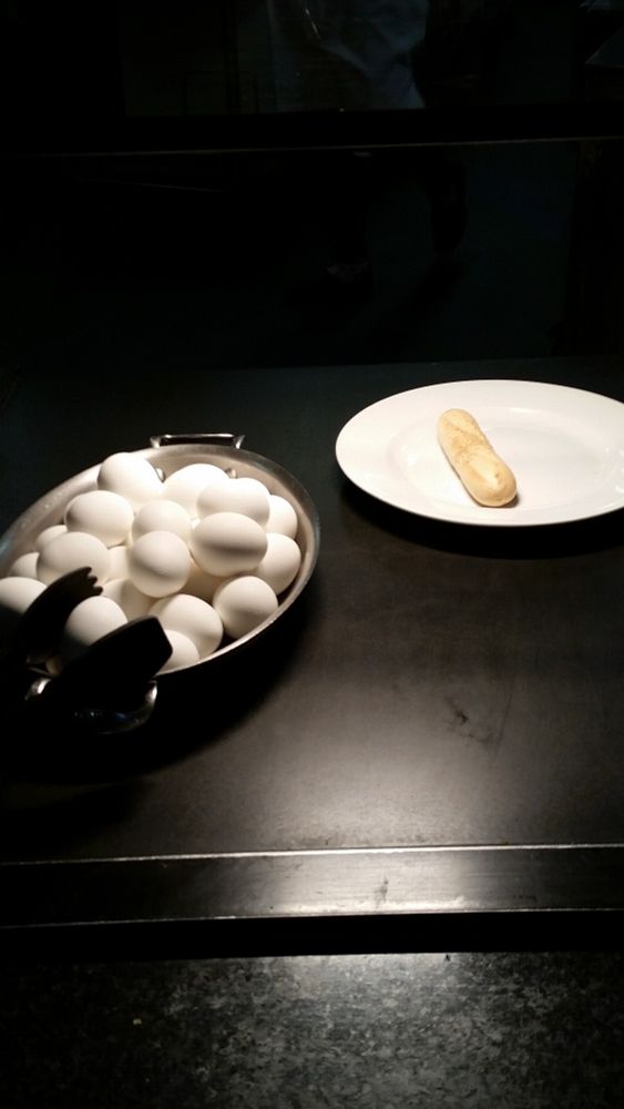 A white dinner plate with a single bread stick sitting on a black surface to the right of a large platter of eggs, one of which is nestled between a pair of tongs.