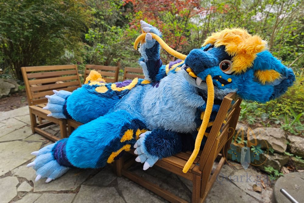 Fursuiter laying on a bench from a lower angle, holding one of the whiskers. Blue noodle dragon with yellow hair and markings
