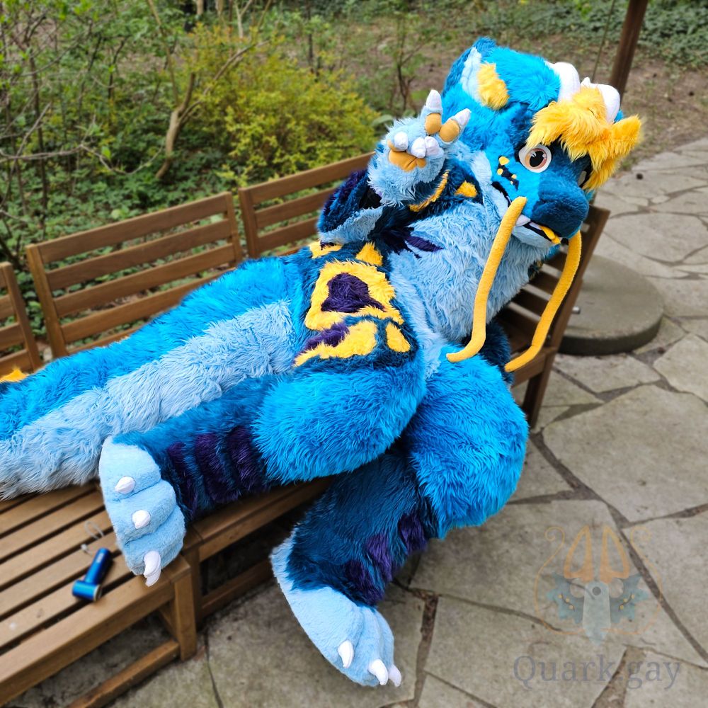 Fursuiter laying down on a bench showing a peace sign. Blue noodle dragon with yellow hair and markings