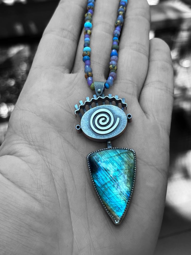 A handmade Sterling silver pendant lays on the palm of someone’s hand. The pendant has a triangular shaped piece of labradorite that hangs down from an abstract idea of what an eye looks like.
