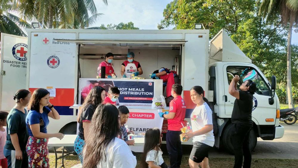 Red Cross volunteers distributing hot meals from a truck to a group of people in a community setting.