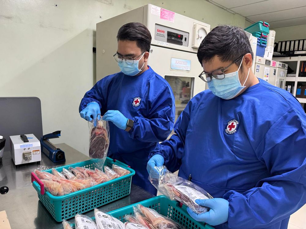 Two Red Cross workers in blue uniforms and masks are handling packaged blood bags in a medical storage room.