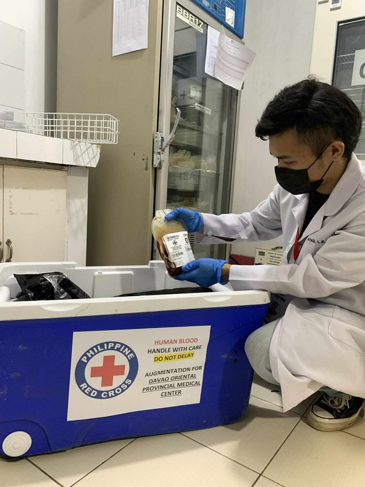A healthcare worker at the Philippine Red Cross is handling a blood donation bag while wearing a lab coat and gloves, sitting next to a cooler marked with the Red Cross logo.