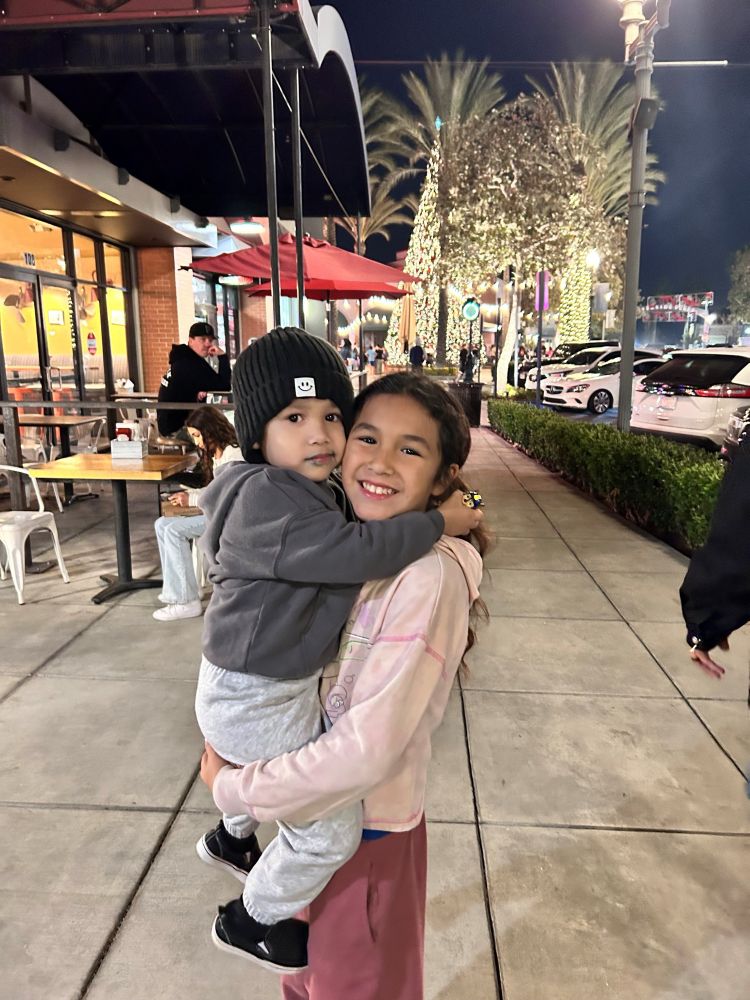 A young girl holding a toddler on a city sidewalk outside of a restaurant with tables on a patio. There are trees with lights around the trunks in the background. 