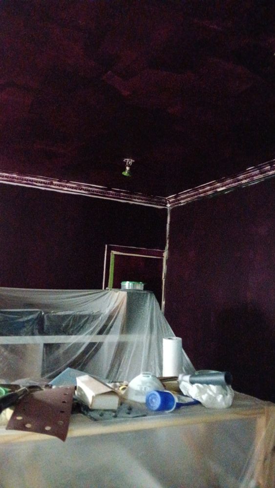 The upper corner of a room mid-painting (the dark purple paint is patchy and there is visible green tape on the edges). In the centre a pile of furniture is covered by dust sheets and has sandpaper and a dust mask on top.