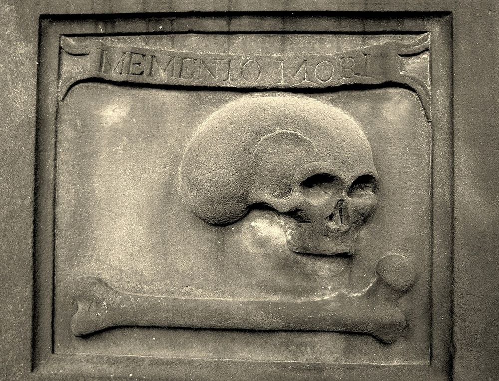 Pierre tombale montrant un crane humain et un os long, surmontés d'une bannière énonçant : Memento Mori