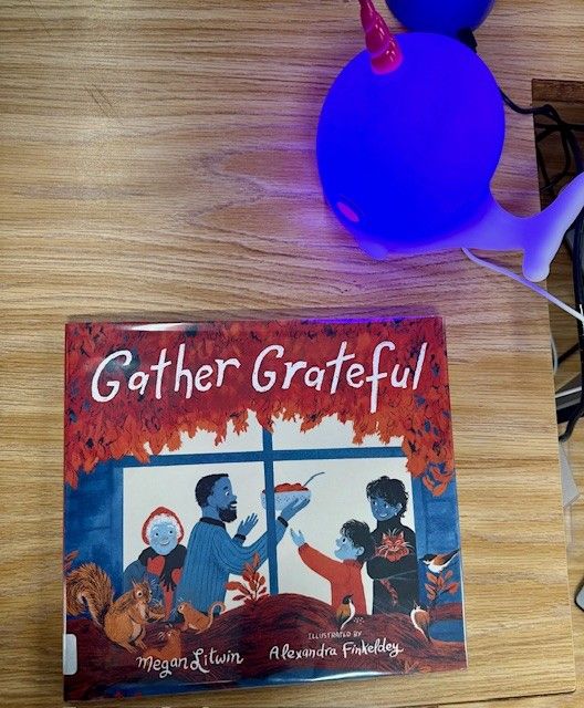 Copy of picture book GATHER GRATEFUL by Megan Litwin with art by Alexandra Finkledey flat on a woodgrain counter. A blue narwhal lamp is on the counter behind the book.
