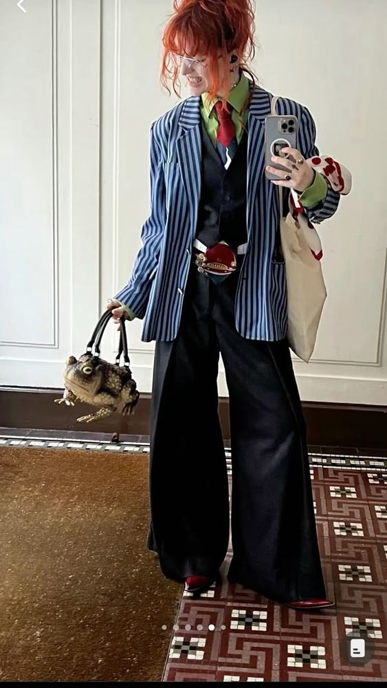 Mulher branca de cabelos laranja usando blazer azul com listras escuras, camisa verde com gravata vermelha por dentro, colete preto, calça preta com barra larga, sapatos vermelhos. Ela está carregando uma ecobag em um braço e uma bolsa em formato de sapo realista no outro.