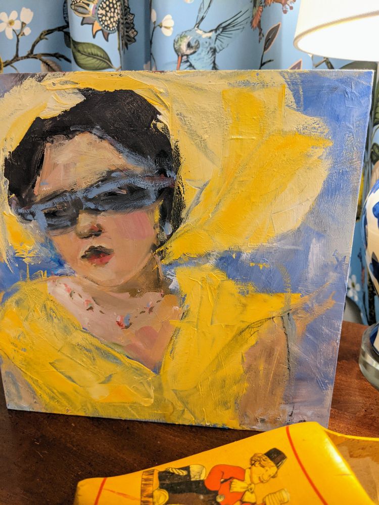 A portrait painting of a woman in a big yellow hat and yellow dress is situated on a table next to a lamp and a yellow wooden shoe.