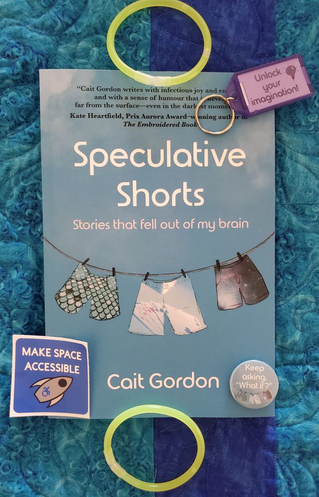 A picture of Speculative Shorts: Stories that fell out of my brain by Cait Gordon. It has a lovely sky-blue cover with three shorts hanging from a line: one covered in scales, one with a pattern of playing cards, and one that shows a starry spacescape. Placed next to the top and bottom of the book are green neon glow bracelets from the CanCon Glow Party - in alignment across from each other. Scattered on and around the book are a sticker that says Make Space Accessible featuring a rocketship, a button featuring the three shorts hanging from a line that are on the book cover and the quote "Keep asking: What if?" and a purple keychain that says "unlock your imagination."